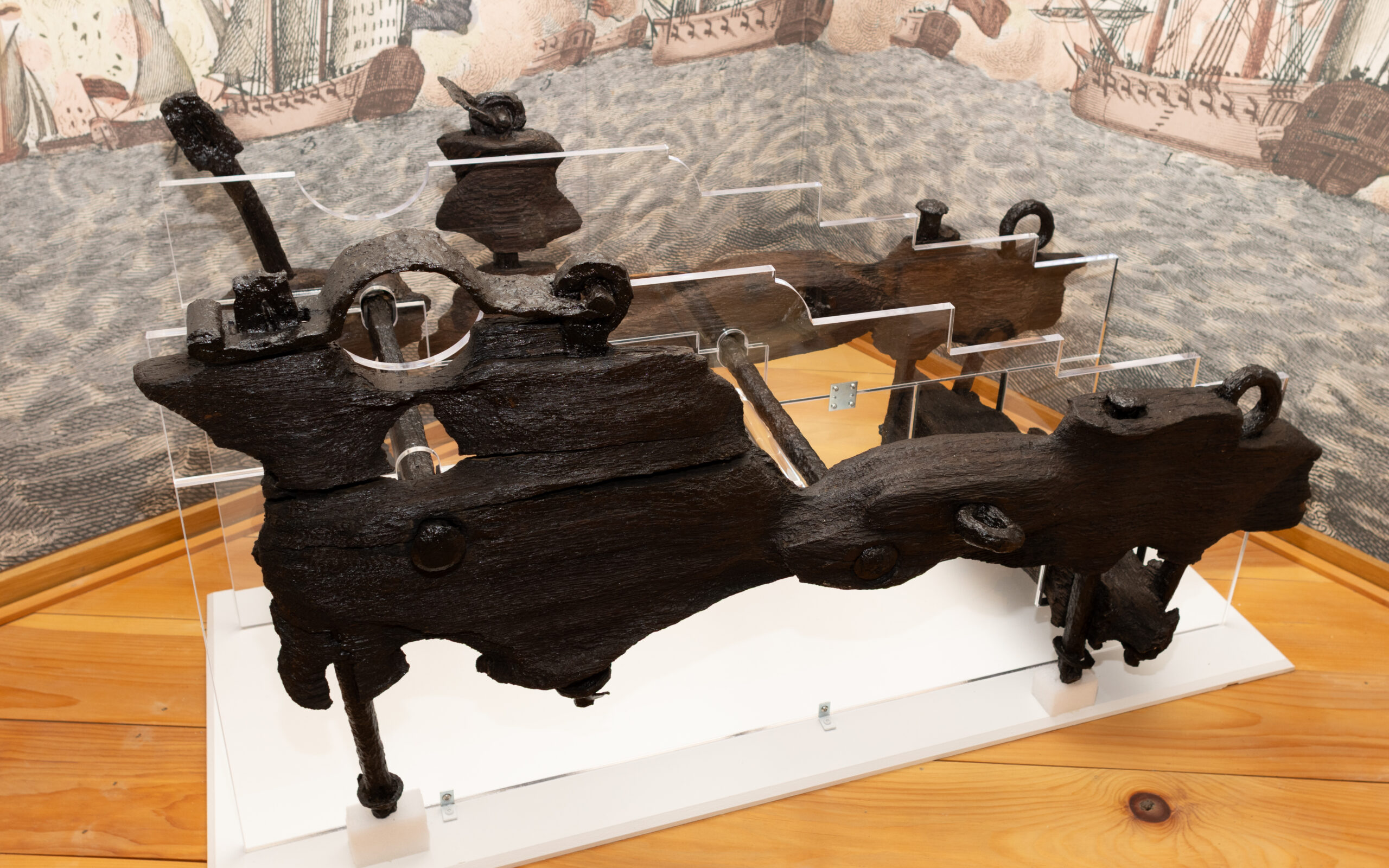 Large dark iron mechanical artifact on a white display base in a museum, with a maritime mural in the background.